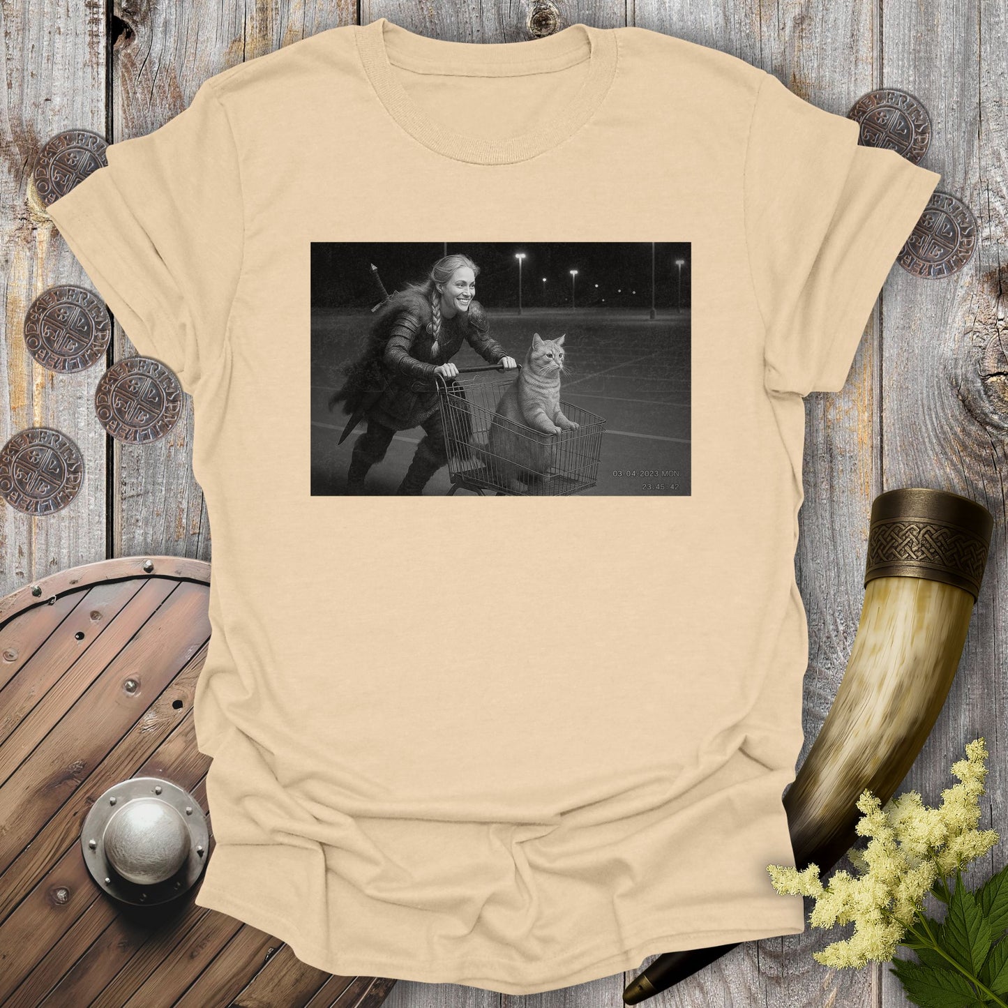 Beige Sand colored t-shirt with a graphic of a Viking Shieldmaiden and a tabby cat in a shopping cart, sprinting on a parking Lot.