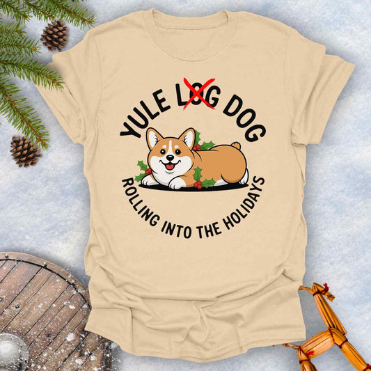 Beige Sand colored T-Shirt with a Corgi holiday graphic and text "Yule Log Dog Rolling Into the Holidays" With "Log" crossed over in red, on a snowy background.