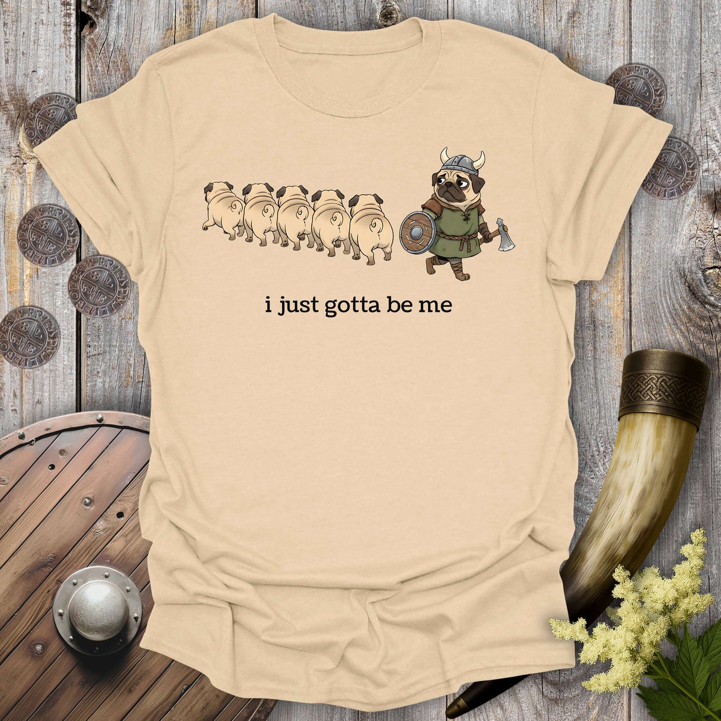 Beige Sand colored t-shirt with cartoon Viking pug design and text "i just gotta be me" on a wooden background.