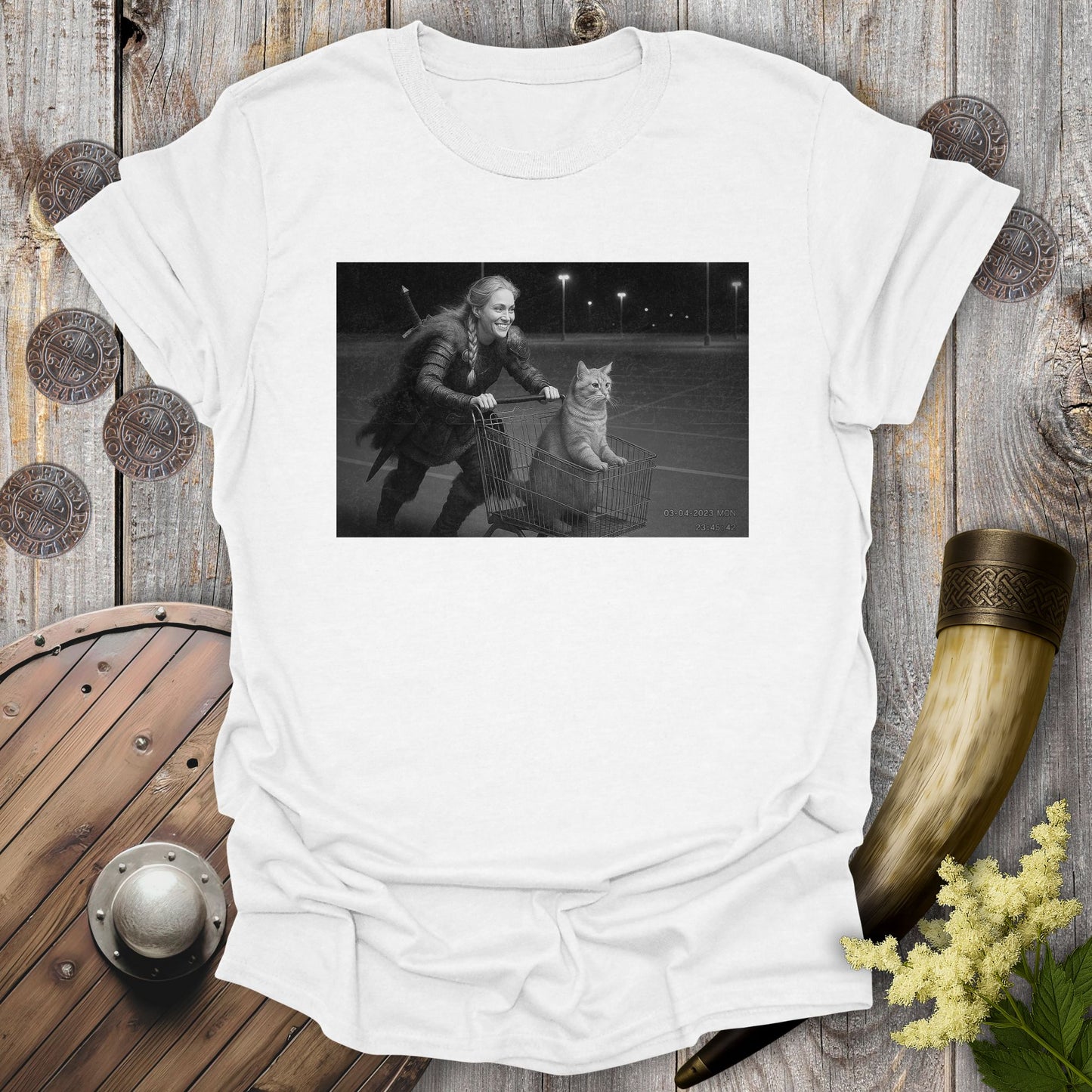 White t-shirt with a graphic of a Viking Shieldmaiden and a tabby cat in a shopping cart, sprinting on a parking Lot.