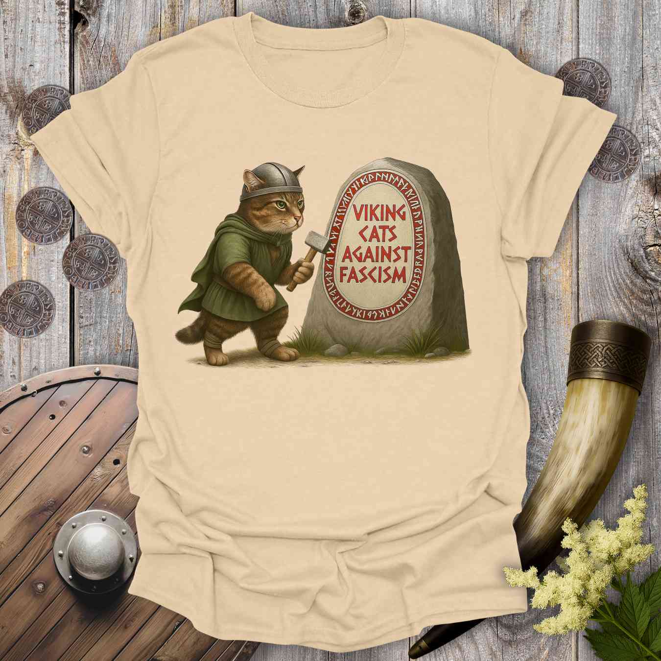 Beige t-shirt with a graphic of a cat in Viking attire and a stone with 'Viking Cats Against Fascism' text, placed on a wooden surface with decorative items.