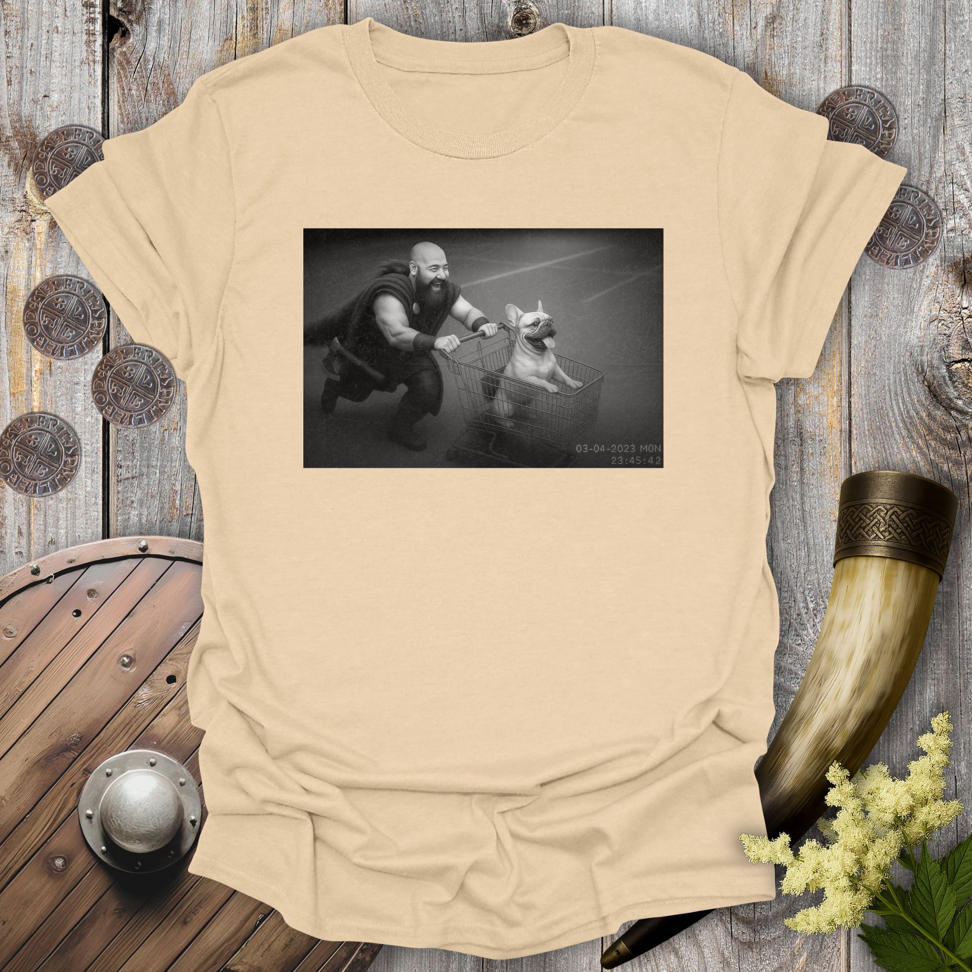 Beige Sand colored t-shirt with a graphic of a Viking and a Bulldog in a shopping cart, sprinting on a parking Lot.