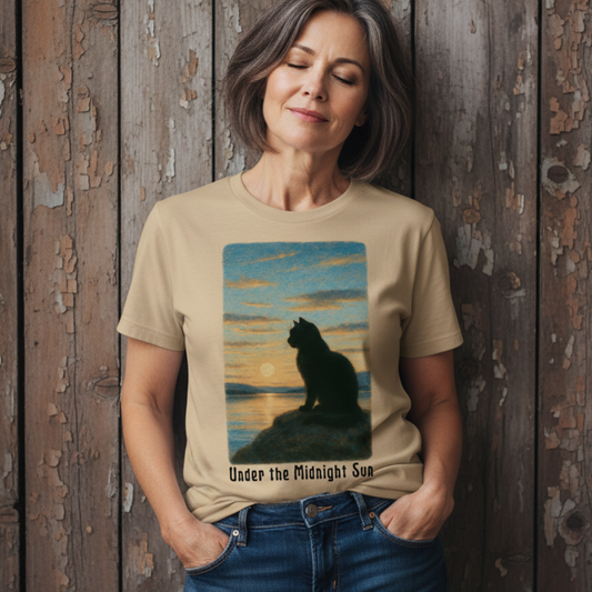 Woman wearing a beige t-shirt with a graphic of a cat and sunset design, with text "Under the Midnight Sun", standing against a wooden wall.