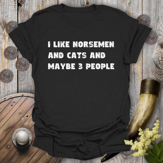 Black t-shirt with white distressed text "I Like Norsemen and Cats and Maybe 3 People" on a wooden background.