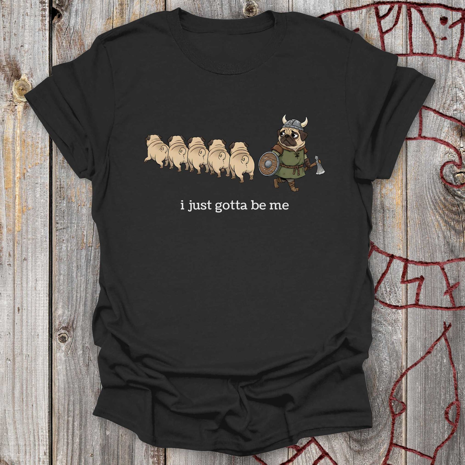 Black t-shirt with cartoon design of a Viking Pug and text 'i just gotta be me' on a wooden surface.