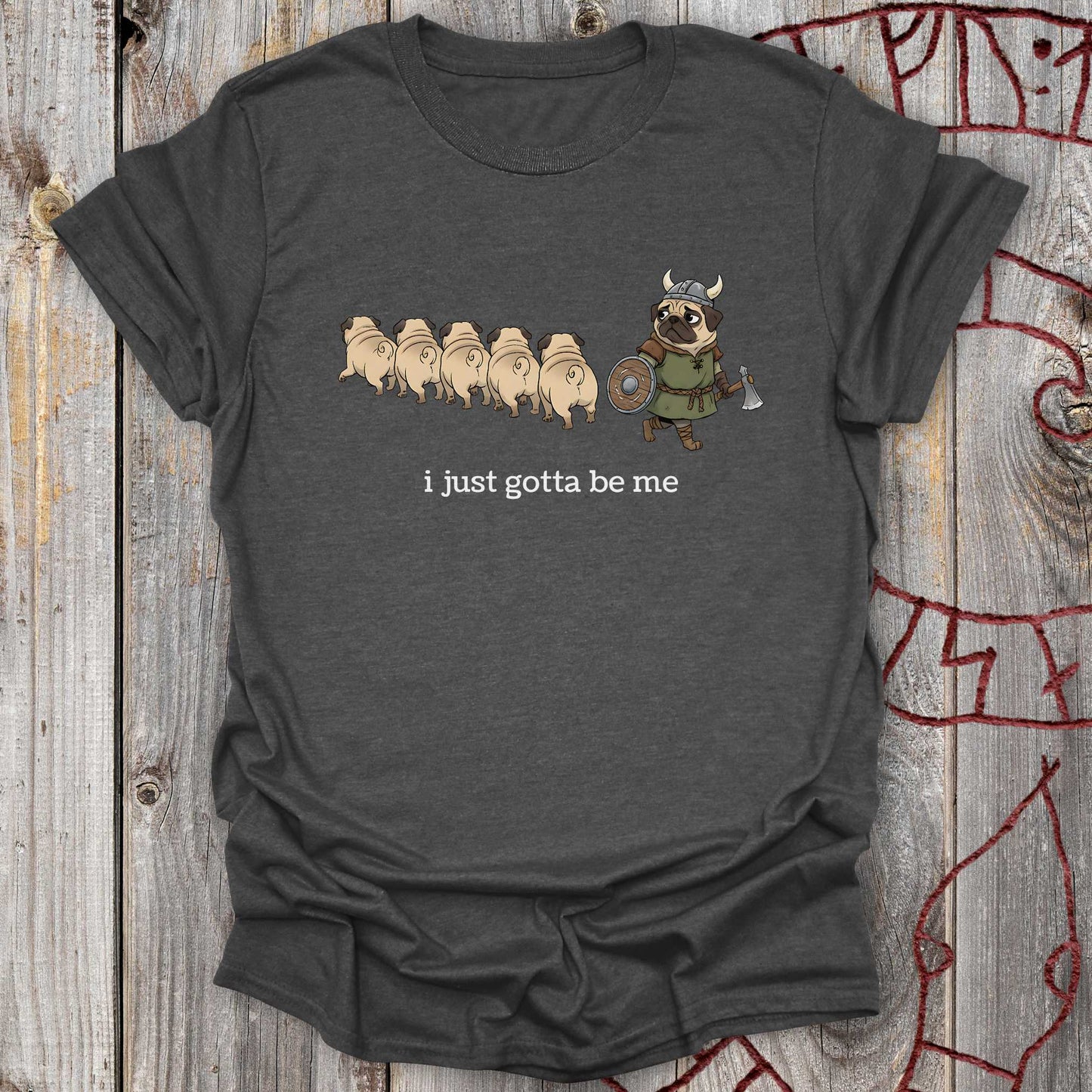 Dark gray t-shirt with cartoon design of a pug in a Viking outfit and text 'i just gotta be me' on a wooden background.