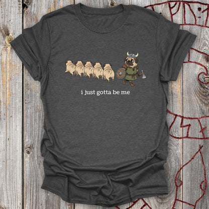 Dark gray t-shirt with cartoon design of a pug in a Viking outfit and text 'i just gotta be me' on a wooden background.