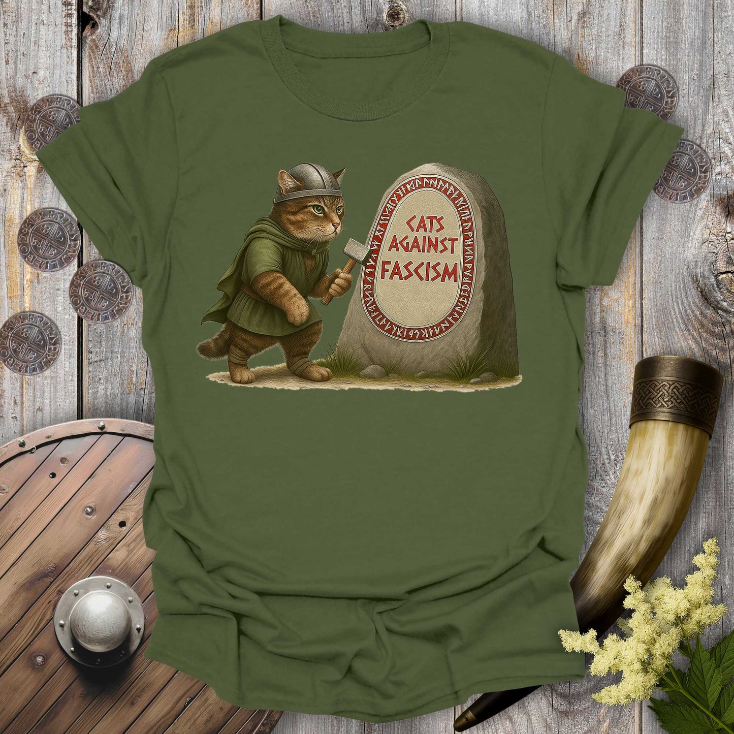 Green t-shirt with a graphic of a cat in Viking attire holding an axe next to a stone with 'Cats Against Fascism' text, displayed on a wooden surface.