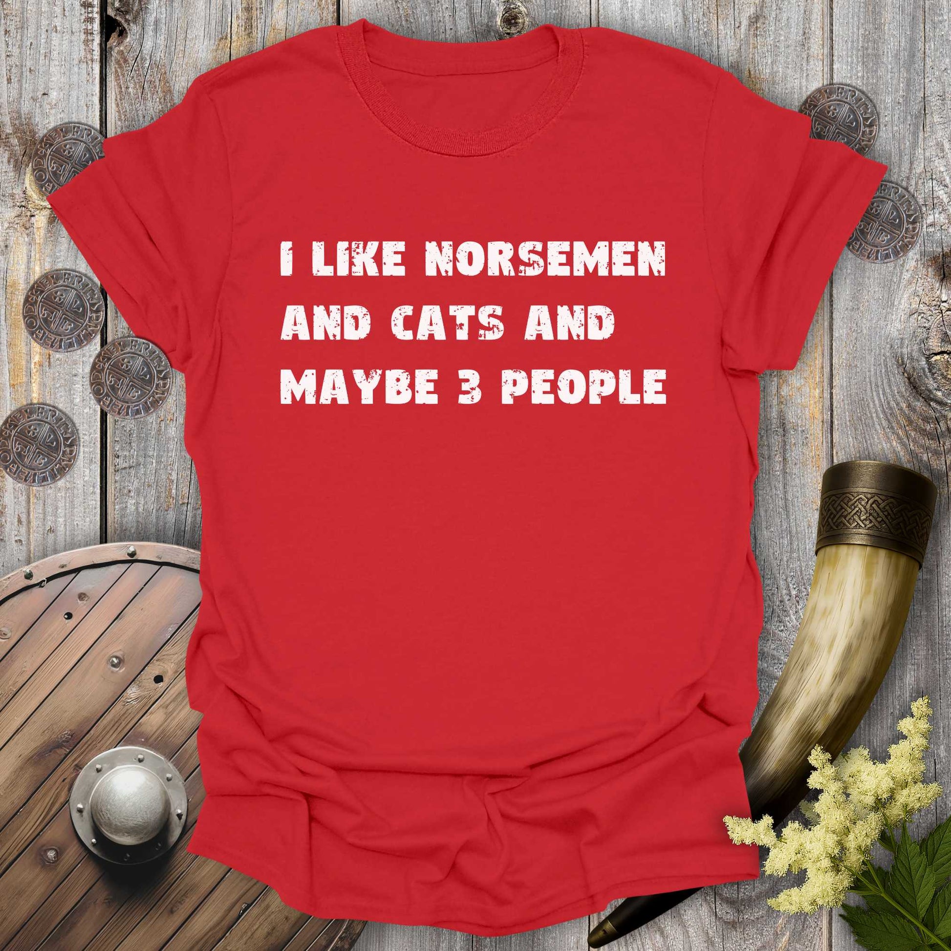 Red t-shirt with white distressed text "I Like Norsemen and Cats and Maybe 3 People" on a wooden background.