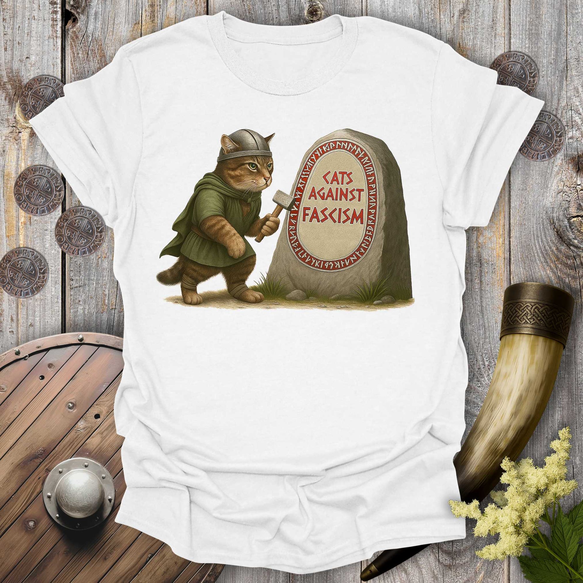 White t-shirt with a graphic of a cat in Viking attire holding an axe next to a stone with 'Cats Against Fascism' text, displayed on a wooden surface.
