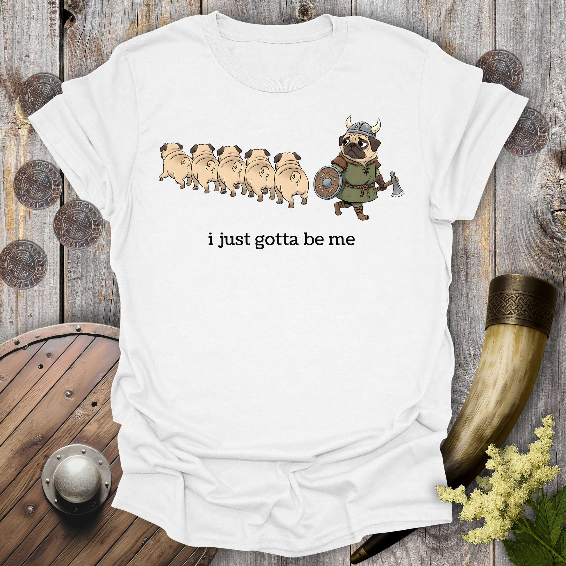 White t-shirt with cartoon Viking pug design and text "i just gotta be me" on a wooden background.