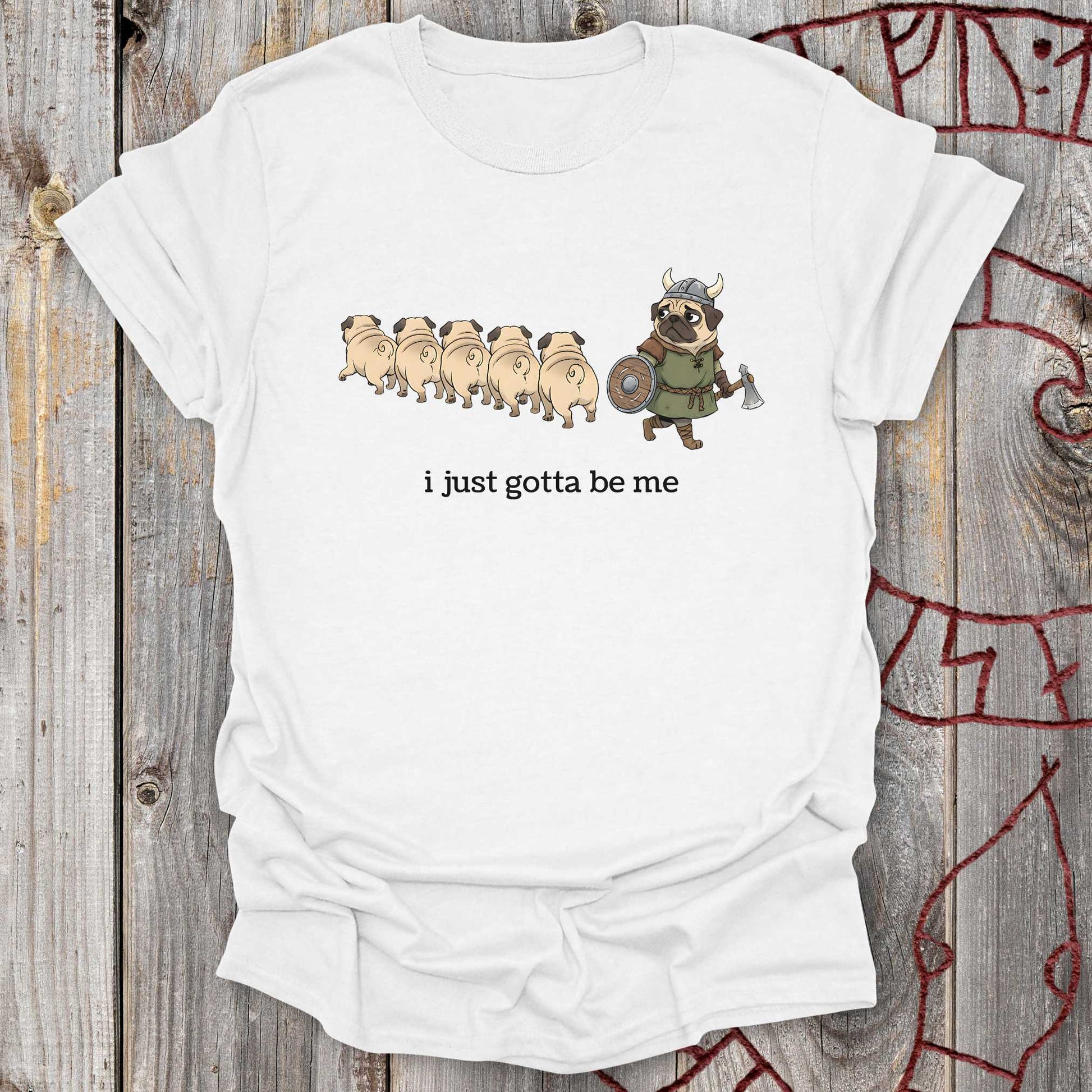 White t-shirt with cartoon design of a pug Viking and text 'i just gotta be me' on a wooden background.