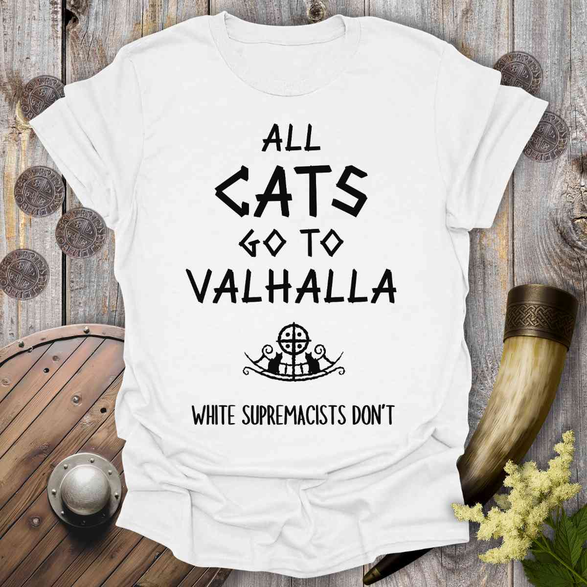 White t-shirt with text 'All cats go to Valhalla, white supremacists don't' on a wooden background.