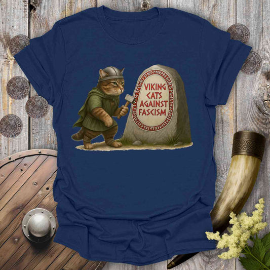 Blue t-shirt with a graphic of a cat in a Viking costume standing next to a stone with 'Viking Cats Against Fascism' text, placed on a wooden background.