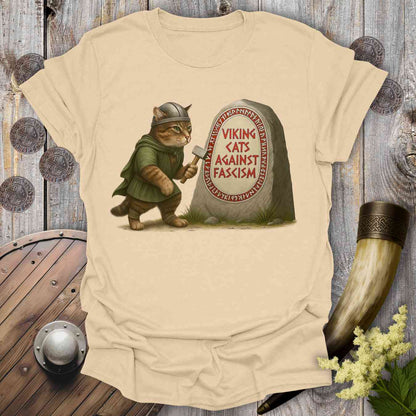 Beige t-shirt with a graphic of a cat in Viking attire and a stone with 'Viking Cats Against Fascism' text, placed on a wooden surface with decorative items.