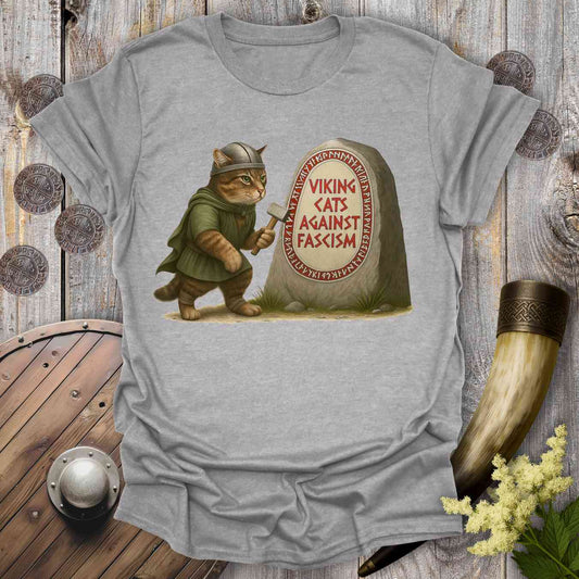 Gray t-shirt with a graphic of a cat in Viking attire next to a stone with 'Viking Cats Against Fascism' text, displayed on a wooden background.