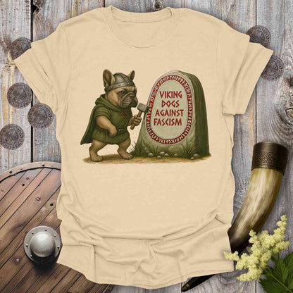 Beige t-shirt with a graphic of a dog in Viking attire holding an axe, standing next to a stone with 'Viking Dogs Against Fascism' text, on a wooden background.