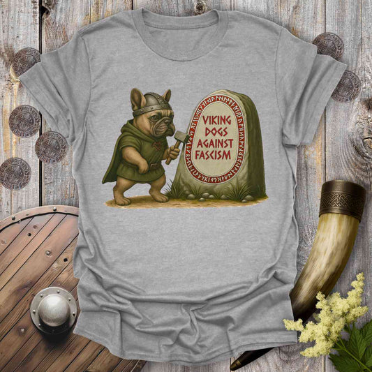 Gray t-shirt with a graphic of a dog in Viking attire holding an ax, standing next to a stone with 'Viking Dogs Against Fascism' text, on a wooden background.